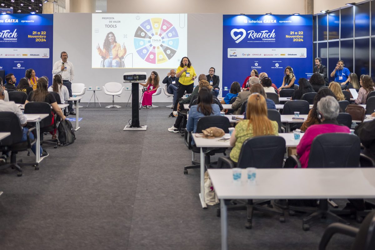 Vista ampla de um painel de discussão sobre ferramentas de inclusão na Reatech. Um palestrante fala ao microfone em um palco baixo, enquanto a plateia assiste sentada em fileiras.