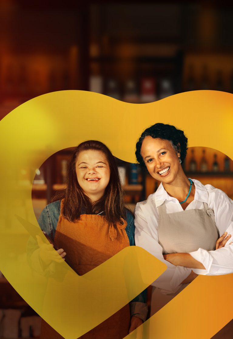 Duas mulheres, uma delas com síndrome de Down, sorriem para a câmera, ambas usando aventais em um ambiente de trabalho.
