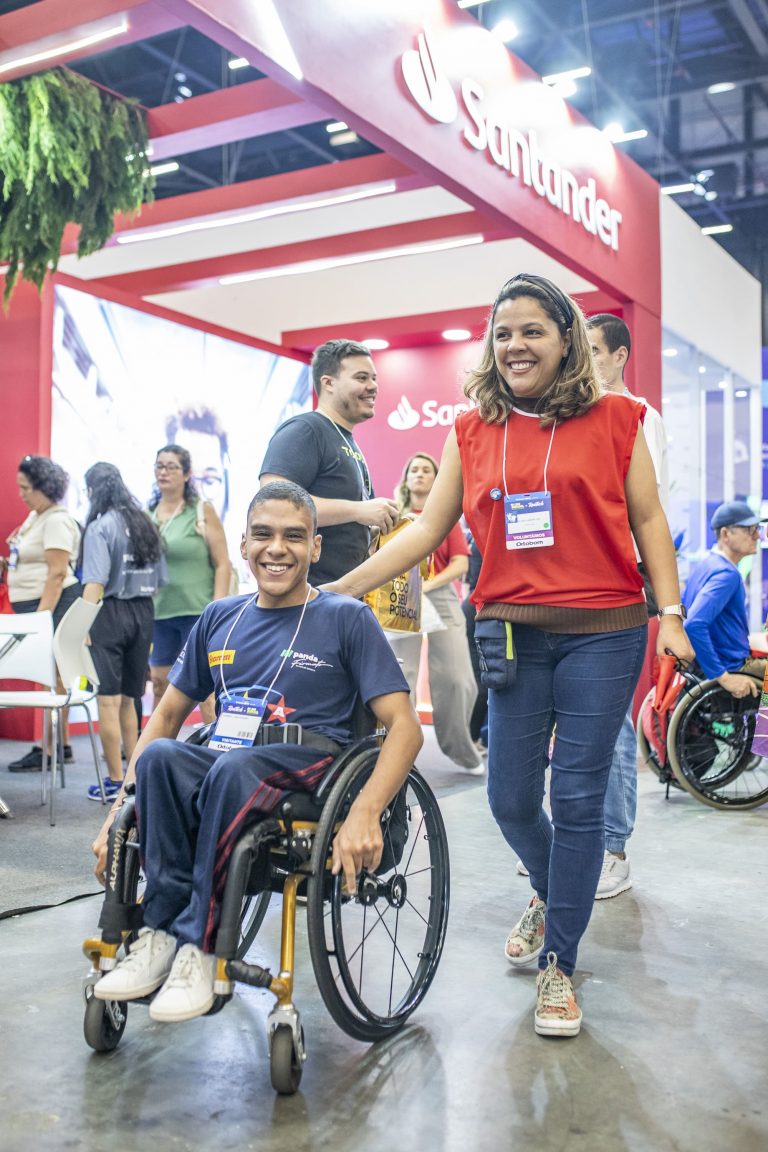 Pessoas no estande do Santander no congresso Reatech. Uma mulher sorridente caminha ao lado de um homem em cadeira de rodas.