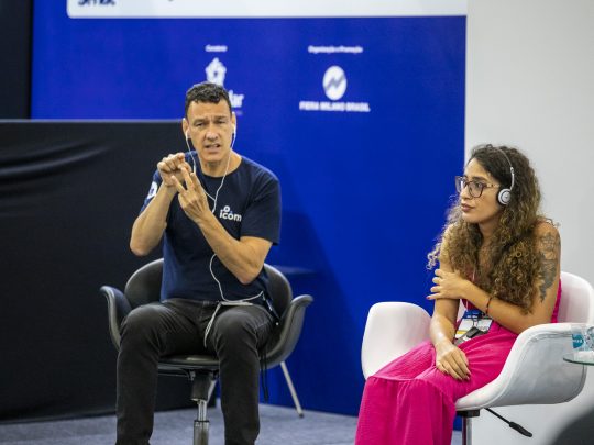 Intérprete de Libras se comunica durante o congresso Reatech. Ao lado dele, uma mulher com fones de ouvido assiste.