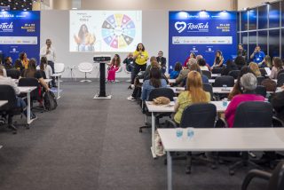 Vista ampla de um painel de discussão sobre ferramentas de inclusão na Reatech. Um palestrante fala ao microfone em um palco baixo, enquanto a plateia assiste sentada em fileiras.