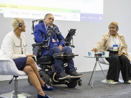 Um homem em cadeira de rodas fala em um painel de discussão com outras duas mulheres no palco do congresso Reatech.